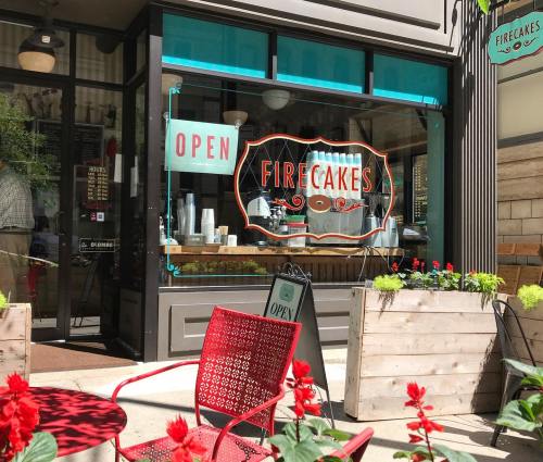 A vibrant storefront of Firecakes with an "Open" sign, surrounded by flowers and outdoor seating.