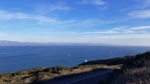 A scenic view of a calm ocean with sailboats, under a clear blue sky and distant mountains on the horizon.