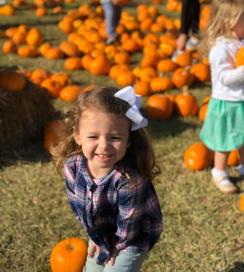 A smiling girl with a bow in her hair stands among pumpkins in a sunny field, with other children in the background.