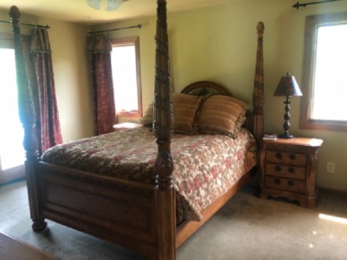 A cozy bedroom featuring a four-poster bed, decorative pillows, a wooden nightstand, and natural light from windows.
