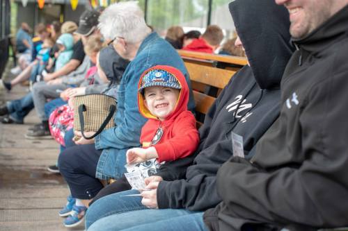 A smiling child in a red hoodie sits on a bench among a group of people, holding a snack and looking cheerful.