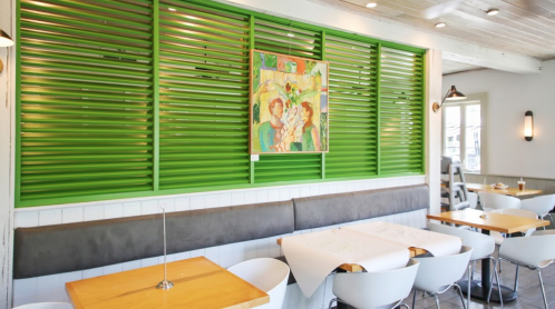 Bright restaurant interior featuring green slatted walls, wooden tables, and a colorful painting.