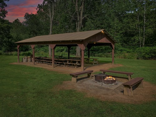 A covered picnic area with benches and a fire pit, surrounded by trees and grass at dusk.