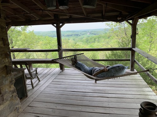 A person relaxes in a hammock on a porch, overlooking a lush green landscape.