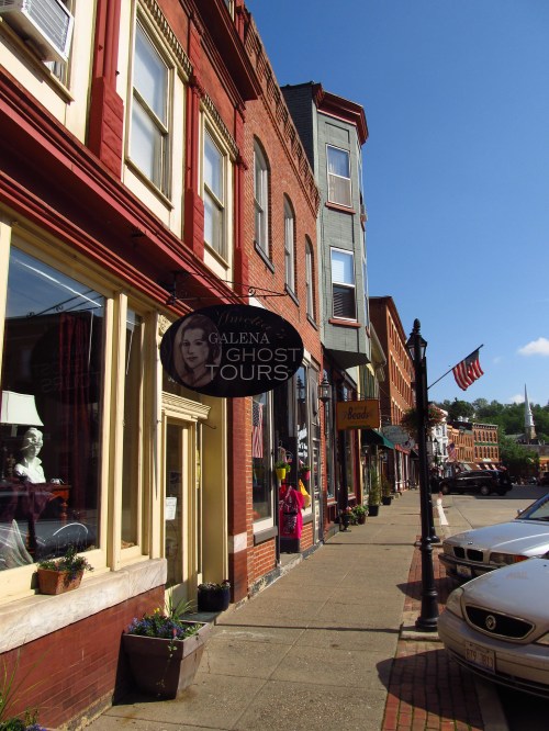 A charming street in Galena featuring historic brick buildings, a ghost tour sign, and American flags.