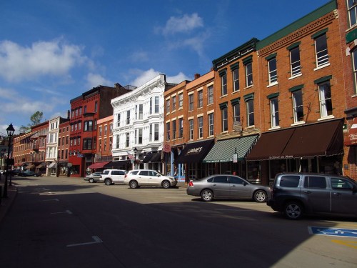 Historic buildings line a street with parked cars under a blue sky and scattered clouds.