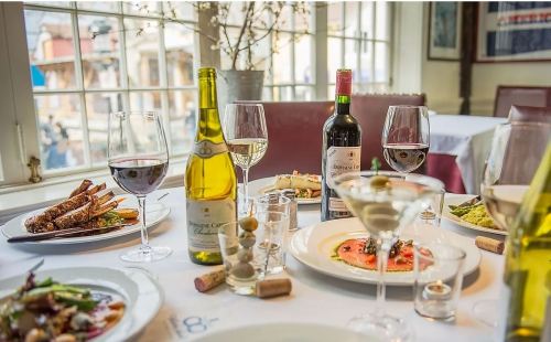 A beautifully set dining table with wine bottles, glasses, and gourmet dishes, featuring elegant decor and natural light.