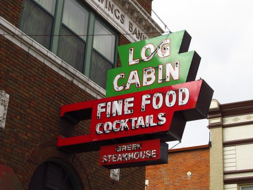 Sign for "Log Cabin" featuring "Fine Food" and "Cocktails," with "Green Steakhouse" below, against a brick wall.