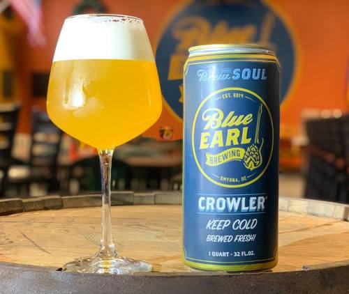 A glass of golden beer next to a can of Blue Earl Brewing Crowler on a wooden barrel in a brewery setting.