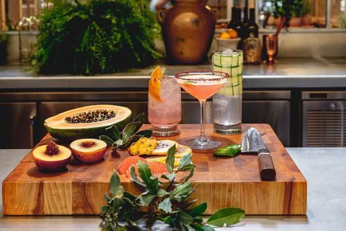 A wooden cutting board displays colorful cocktails and fresh fruits, including papaya, peaches, and grapefruit, with a knife.