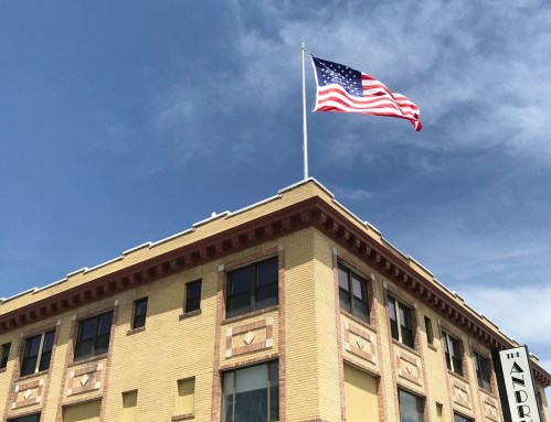 A large American flag waves atop a historic brick building under a blue sky with scattered clouds.