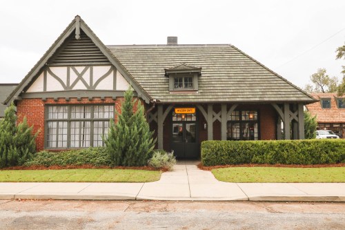 A charming brick house with a green roof and decorative trim, surrounded by neatly trimmed hedges and a sidewalk.