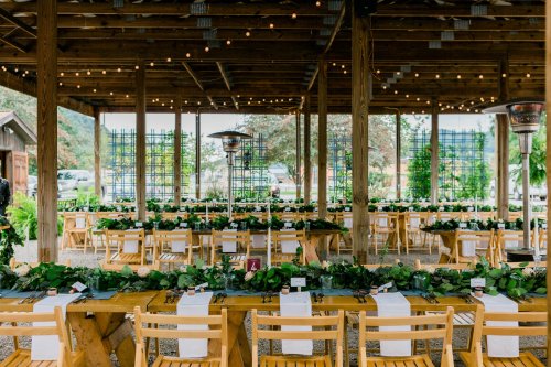 A rustic outdoor dining setup with wooden tables, greenery, and string lights under a wooden structure.