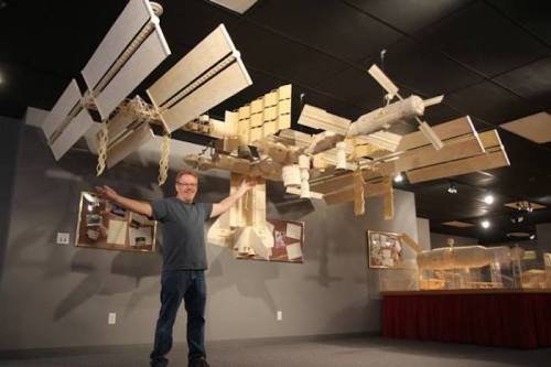 A man stands with arms outstretched beside a large model of the International Space Station in a museum.