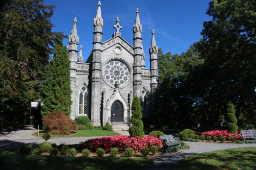 Gothic-style church with a rose window, surrounded by greenery and colorful flowers under a clear blue sky.