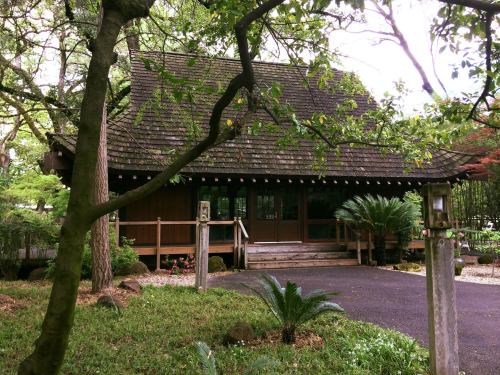 A traditional wooden house with a sloped roof, surrounded by greenery and trees in a serene garden setting.