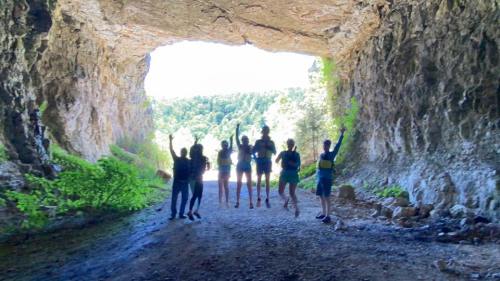 A group of people joyfully jumping in a cave entrance, surrounded by greenery and bright sunlight.