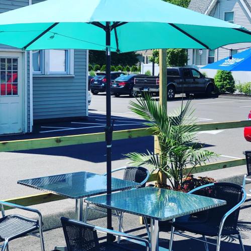 A turquoise umbrella shades a table and chairs in a parking lot, with a palm plant nearby and cars in the background.