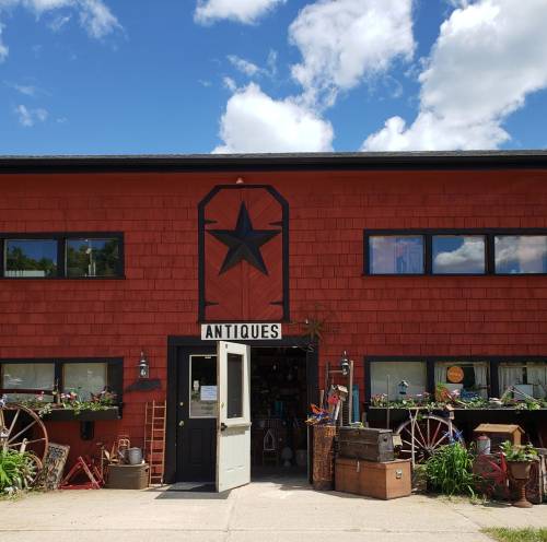 A red antique shop with a black star, large windows, and a welcoming entrance surrounded by greenery and vintage items.