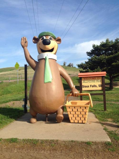 A large bear statue wearing a green hat and scarf waves next to a picnic basket, welcoming visitors to Jellystone Park.