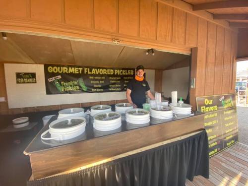 A vendor stands behind a table displaying various gourmet pickles in white buckets, with a sign listing flavors.