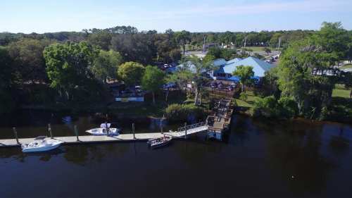 Aerial view of a riverside restaurant with boats docked, surrounded by trees and a clear blue sky.
