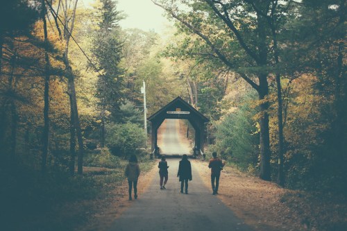 A group of people walking towards a covered bridge surrounded by autumn trees and foliage.