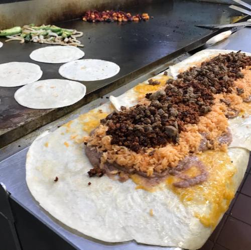 A large tortilla filled with rice, beans, and seasoned meat, with tortillas cooking on a griddle in the background.
