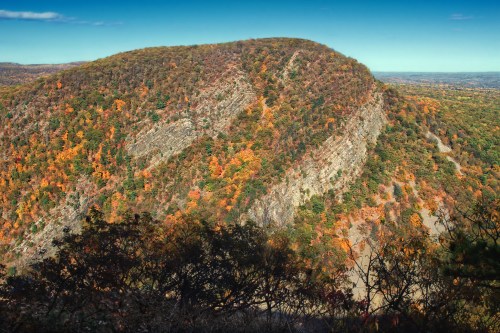 A scenic view of a mountain covered in autumn foliage, showcasing vibrant orange and green trees against a clear blue sky.