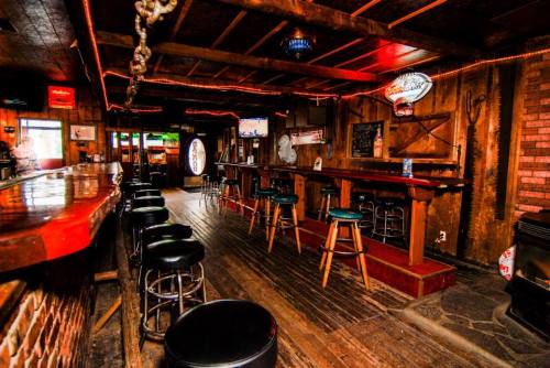 A rustic bar interior with wooden beams, stools, and dim lighting, featuring a long counter and a cozy atmosphere.