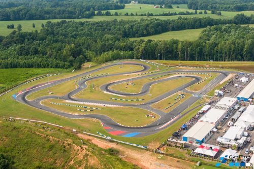 Aerial view of a go-kart track surrounded by green fields and trees, with a nearby event area and tents.