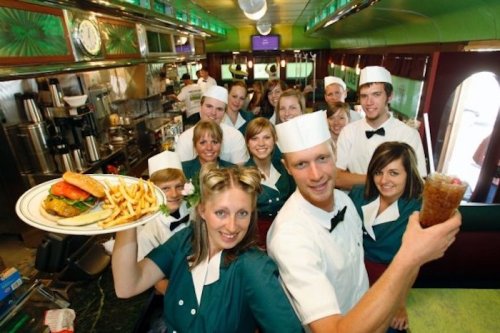 A group of smiling diner staff in retro uniforms, holding food and drinks, posing together in a vibrant diner setting.