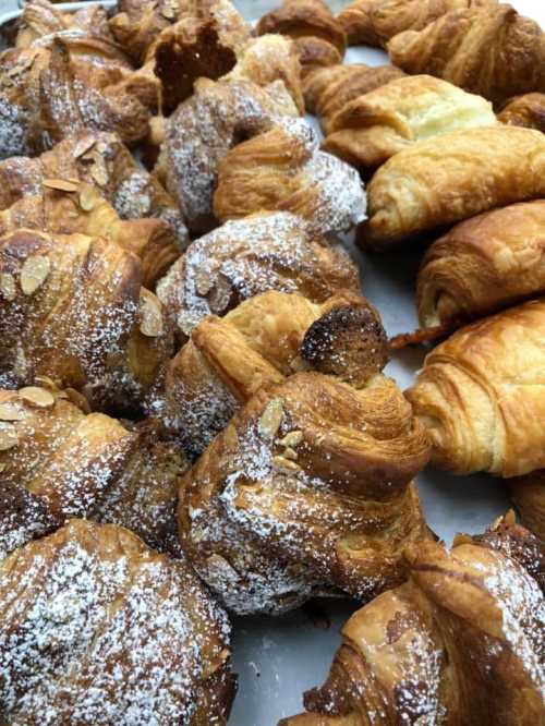 A variety of freshly baked pastries, including croissants and almond croissants, dusted with powdered sugar.