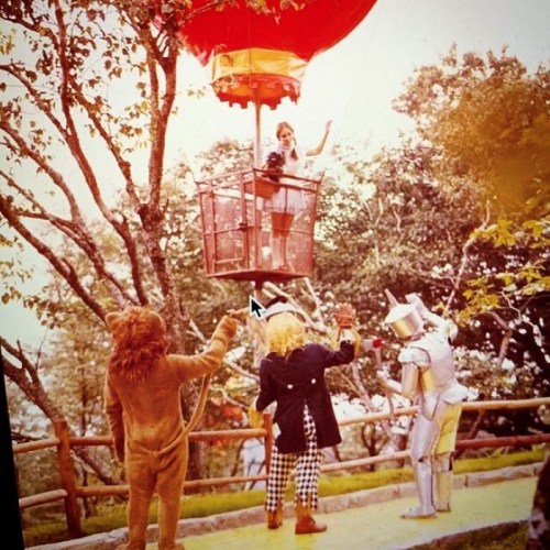 A vintage scene featuring a girl in a hot air balloon with a lion, scarecrow, and tin man below in a whimsical setting.