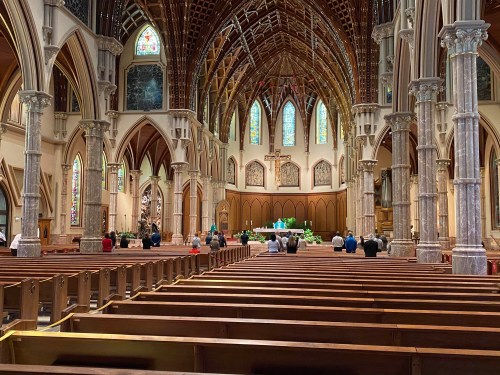 Interior of a grand church with tall arches, stained glass windows, and people seated in wooden pews.