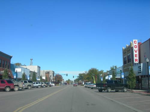 A view of a downtown street with shops, parked cars, and a clear blue sky.