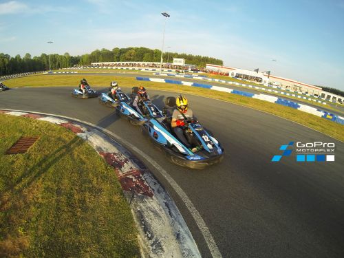 Four go-karts racing on a track at GoPro Motorplex, with a clear blue sky and green trees in the background.