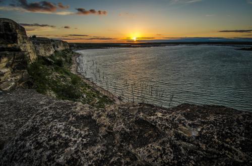 Sunset over a calm lake, with rocky cliffs and lush greenery in the foreground, casting warm colors across the sky.
