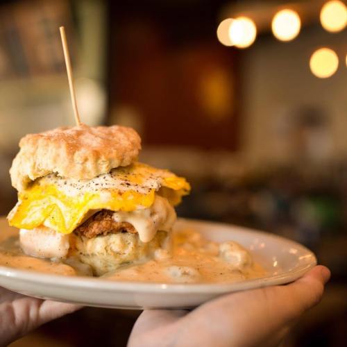 A hand holds a plate with a biscuit sandwich topped with fried egg and gravy, set in a cozy restaurant atmosphere.