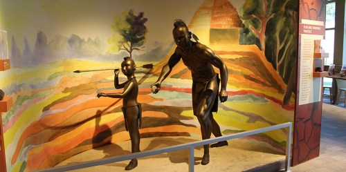 A bronze sculpture of a man and child, set against a colorful mural of a landscape, depicting a cultural scene.