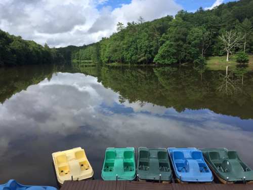 Colorful paddle boats lined up on a dock, reflecting in a calm lake surrounded by lush green trees and a cloudy sky.