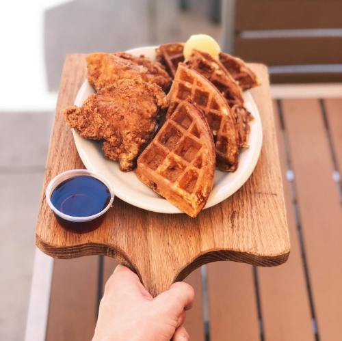 A hand holding a wooden platter with fried chicken, waffles, and a small cup of syrup.