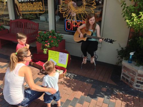 A young woman plays guitar while seated outside, as a woman and two children listen nearby. Bright flowers and an "Open" sign are visible.