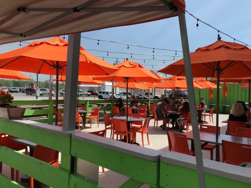 Outdoor dining area with orange umbrellas, green walls, and patrons enjoying their meals under string lights.