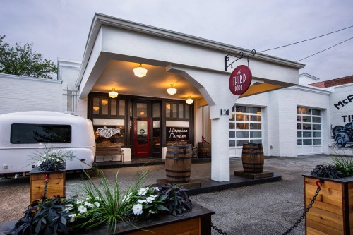 Exterior of a white building with a sign reading "The Third" and barrels, featuring a cozy entrance and plants.