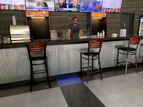 A man stands behind a bar with metal panels, featuring several drink taps and empty stools in a casual setting.
