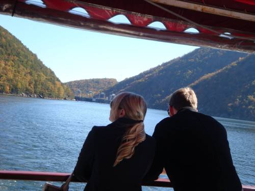 A couple stands on a boat, gazing at a scenic river surrounded by autumn-colored hills.