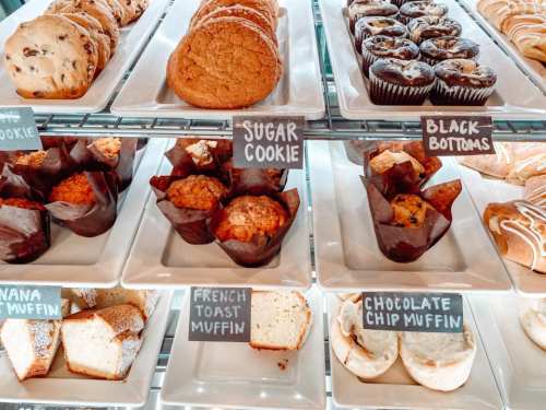 A display of assorted baked goods, including cookies, muffins, and cupcakes, on white plates with labels.