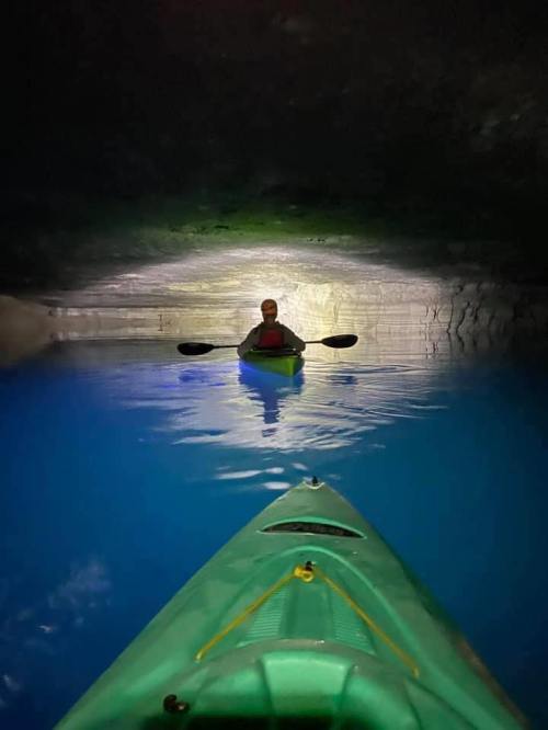 A person in a kayak paddles through a dark cave with glowing blue water and illuminated rock walls.