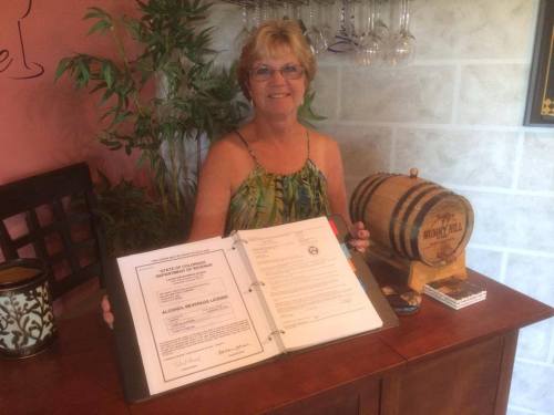 A woman smiles while holding an open binder with an alcohol license, next to a small barrel and decorative items.